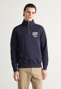 Hanorac GANT bleumarin, cu guler înalt, fermoar pe jumătate în față și detalii ale logo-ului alb pe piept. Fabrică din bumbac moale și talie robustă.