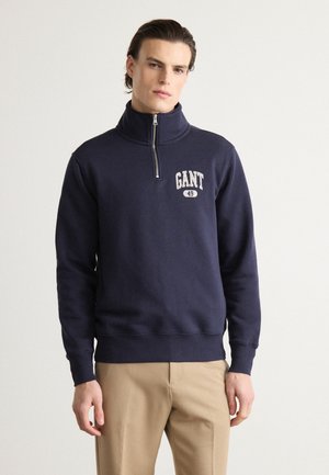 Mornarnički plavi GANT dukserica s visokim ovratnikom, polu-zip prednjim dijelom i bijelim logotipom na prsima. Mekana pamučna tkanina i čvrsta elastična traka.