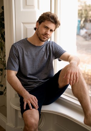 Grijze T-shirt met korte mouwen van zacht materiaal, gecombineerd met donkere shorts. Het onderwerp zit bij een raam in een ontspannen houding.