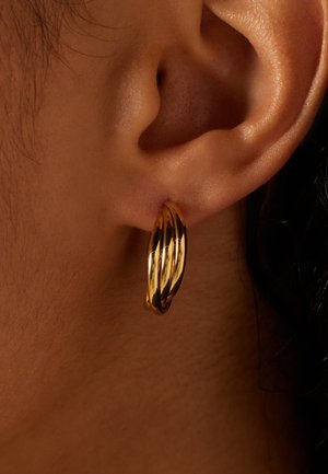 Goldene Creolen mit einem gedrehten Design, polierte Oberfläche, mittlere Größe, am Ohrläppchen befestigt. Texturierte Haut im Hintergrund sichtbar.