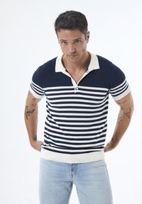 Mørkeblå og hvid stribet polo skjorte med ribbede kraver og manchetter. Korte ærmer, tætsiddende design og knaplukning ved halsen.