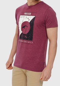 Maglietta maroon in tessuto morbido, con una stampa grafica con accenti neri, design circolare e il testo "INDICODE JEANSPIRIT" in alto.