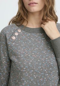 Sudadera gris con un patrón disperso de puntos rosas, azules y blancos. Incluye tres botones rosas en el hombro y puños acanalados.