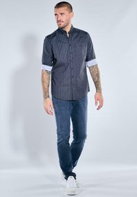 Marineblau gemustertes Hemd mit kurzen Ärmeln und weißen Manschetten, kombiniert mit dunkelblauen Jeans und weißen Turnschuhen. Modell steht lässig.