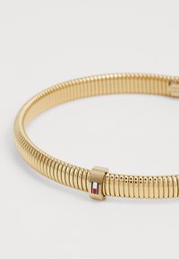 Collier ras du cou doré avec un design flexible et rainuré, et un petit accent logo aux couleurs rouge, blanc et bleu.