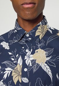 Camicia blu navy con motivi floreali bianchi e beige, colletto standard e chiusura con bottoni, tessuto morbido e texture liscia.