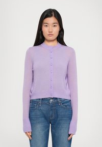 Vaaleanlila lyhyt neulecardigan, jossa on pyöreä kaulus, pitkät hihat ja nappikiinnitys edessä. Ribattuja hiha- ja helmareunoja, jotka lisäävät tekstuuria. Viety denim-farkkujen päälle.