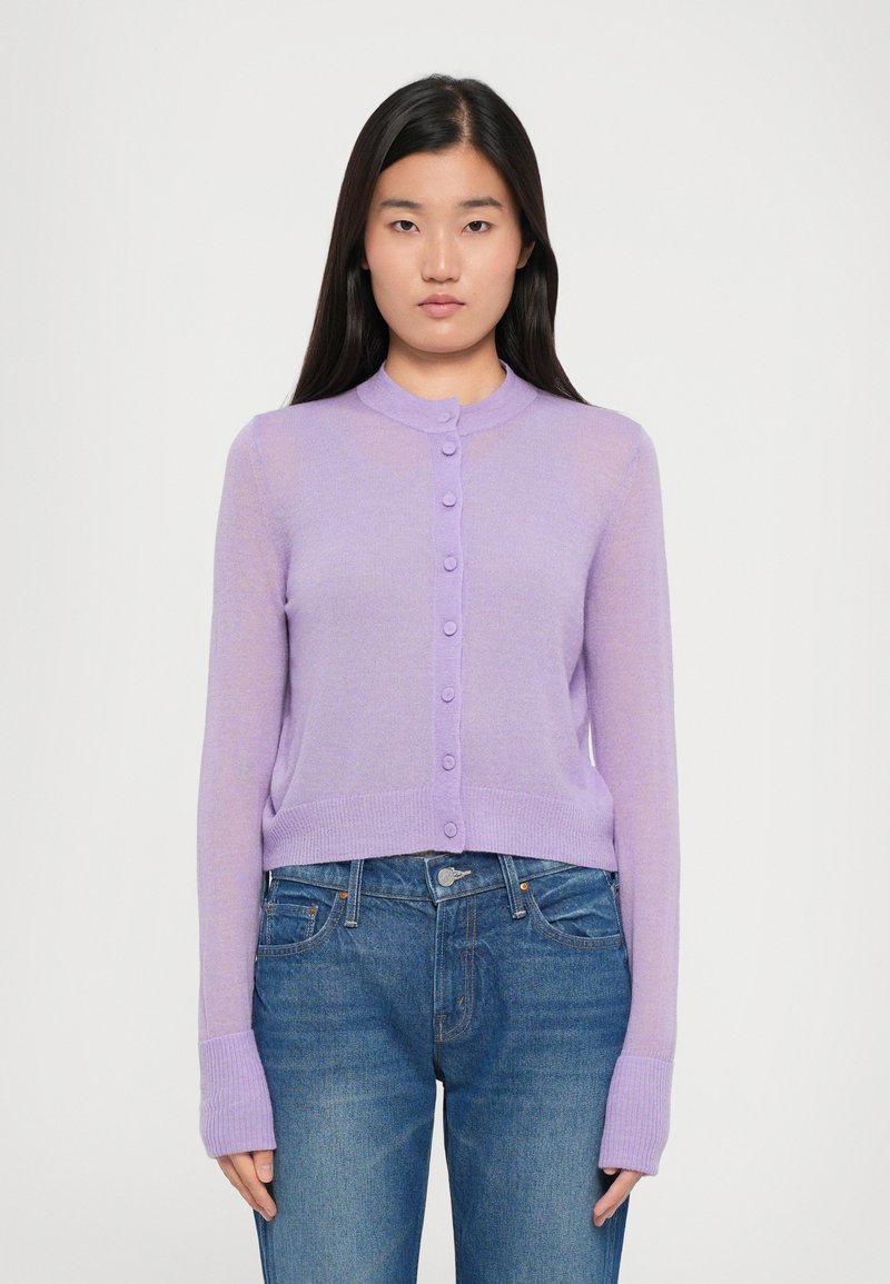 Vaaleanlila lyhyt neulecardigan, jossa on pyöreä kaulus, pitkät hihat ja nappikiinnitys edessä. Ribattuja hiha- ja helmareunoja, jotka lisäävät tekstuuria. Viety denim-farkkujen päälle.