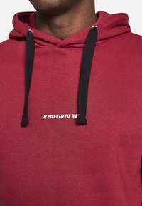 Červená mikina s kapucí vyrobená z měkké tkaniny, s černými šňůrkami a vytištěným textem "REDEFINED RED" v bílé barvě. Jednoduchý, ležérní design.