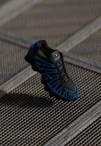 Sapatilha desportiva preta e azul com sola em forma de mola repousa sobre uma superfície de grelha metálica texturizada.