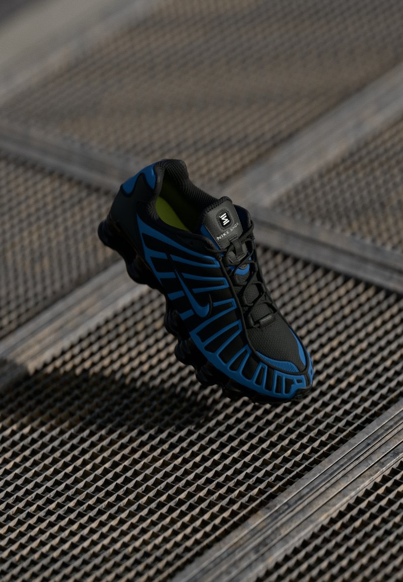 Sapatilha desportiva preta e azul com sola em forma de mola repousa sobre uma superfície de grelha metálica texturizada.