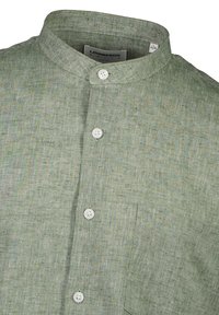 Camicia di lino in verde tenue, con colletto a bandana, bottoni bianchi e una tasca a toppa, che mette in mostra una trama tessuta.