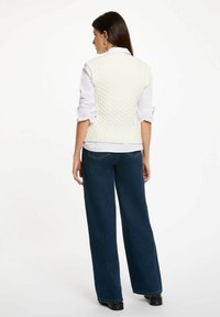 Gilet bianco lavorato a maglia con motivo testurizzato, scollatura rotonda e orlo a coste, indossato sopra una camicia bianca e abbinato a jeans larghi scuri.