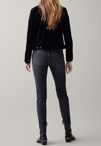 Musta samettikuvioinen takki napituksella, yhdistetty tummanharmaisiin slim-fit-farkkuihin ja mustiin saappaisiin, kuvattuna takaa neutraalia taustaa vasten.