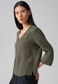 Blusa verde oliva, con texture, caratterizzata da scollatura a V, chiusura con bottoni e maniche a tre quarti, con uno spacco laterale per un tocco di dettaglio.