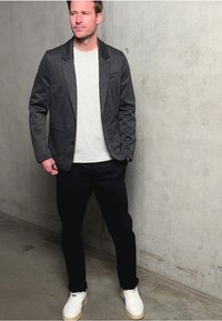 Harmaa bleiseri vaaleanharmaan t-paidan, tummien housujen ja valkoisten lenkkareiden päällä. Taustalla betoni seinä. Bleiserissä on hammastettu kaulus ja yksi nappi.