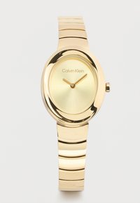 Χρυσό ρολόι Calvin Klein με οβάλ καντράν, μινιμαλιστικό καντράν και μεταλλικό βραχιόλι με συνδέσμους σε λευκό φόντο.