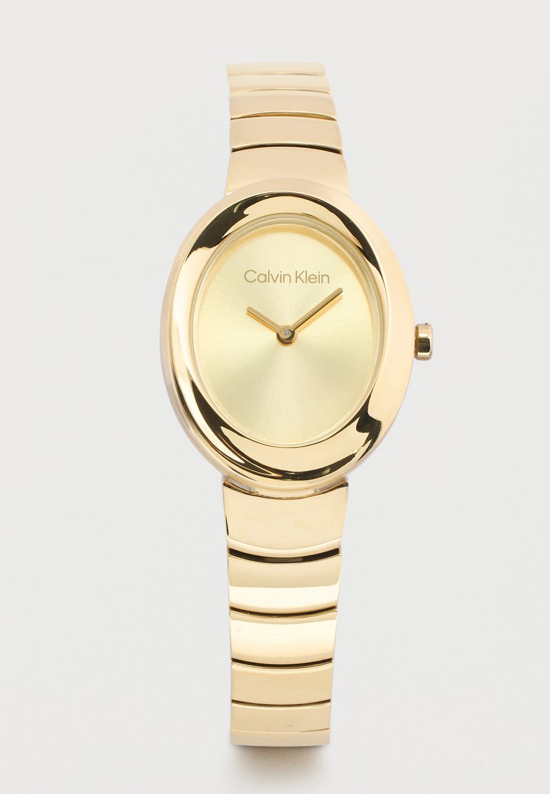 Χρυσό ρολόι Calvin Klein με οβάλ καντράν, μινιμαλιστικό καντράν και μεταλλικό βραχιόλι με συνδέσμους σε λευκό φόντο.