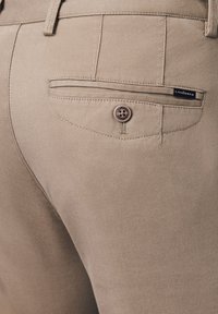 Beżowe spodnie o gładkiej fakturze, z tylną kieszenią zapinaną na guzik i czarną metką na krawędzi kieszeni.