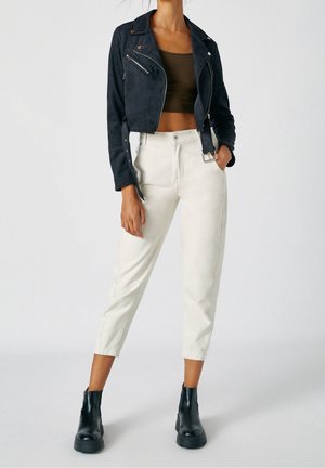 Kobieta w czarnej skróconej kurtce motocyklowej, ciemnobrązowym krótkim topie, kremowych spodniach z wysokim stanem i czarnych botkach do kostki, stojąca z jedną ręką w kieszeni.