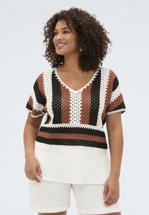 Kvinde smilende iført en kortærmet hæklet top med brune, sorte og hvide striber over en hvid bluse og hvide shorts.