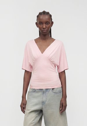 Mujer con el cabello trenzado que lleva una camiseta rosa pálido con cuello en V y mangas cortas y vaqueros anchos de lavado claro, de pie contra un fondo liso.