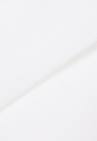 Valkoinen kuvioitu kangas taitettuna diagonaalisesti, paljastaen hienovaraisia kuvioita ja pehmeitä varjoja yksinkertaisella taustalla.