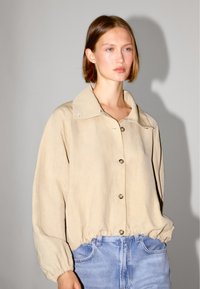 Beżowa, lekka kurtka z dużym kołnierzem, zapinana na guziki z elastycznym dołem. Charakteryzuje się miękką fakturą i subtelnymi detalami przeszyć.