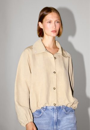 Beige, let jakke med stor krave, knaplukning foran og elastisk kant. Har blød tekstur og diskrete syningsdetaljer.