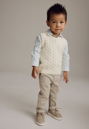 Jeune garçon debout portant un gilet en maille torsadée crème, une chemise rayée à boutons, un pantalon beige et des chaussures en daim beige sur un fond uni.