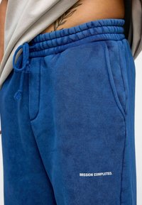 Blauwe fleece joggingbroeken met een elastische tailleband, zijzakken en een trekkoord. Bevat de tekst "MISSIE VOLTOOID." in het wit.