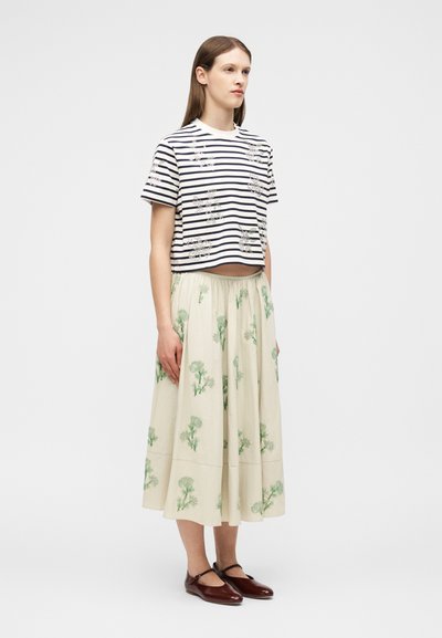 Γυναίκα με ίσια καστανά μαλλιά, φοράει μαύρο και λευκό ριγέ κοντομάνικο τοπ και κρεμ φούστα με πράσινα ανθισμένα σχέδια, στέκεται ακίνητη.