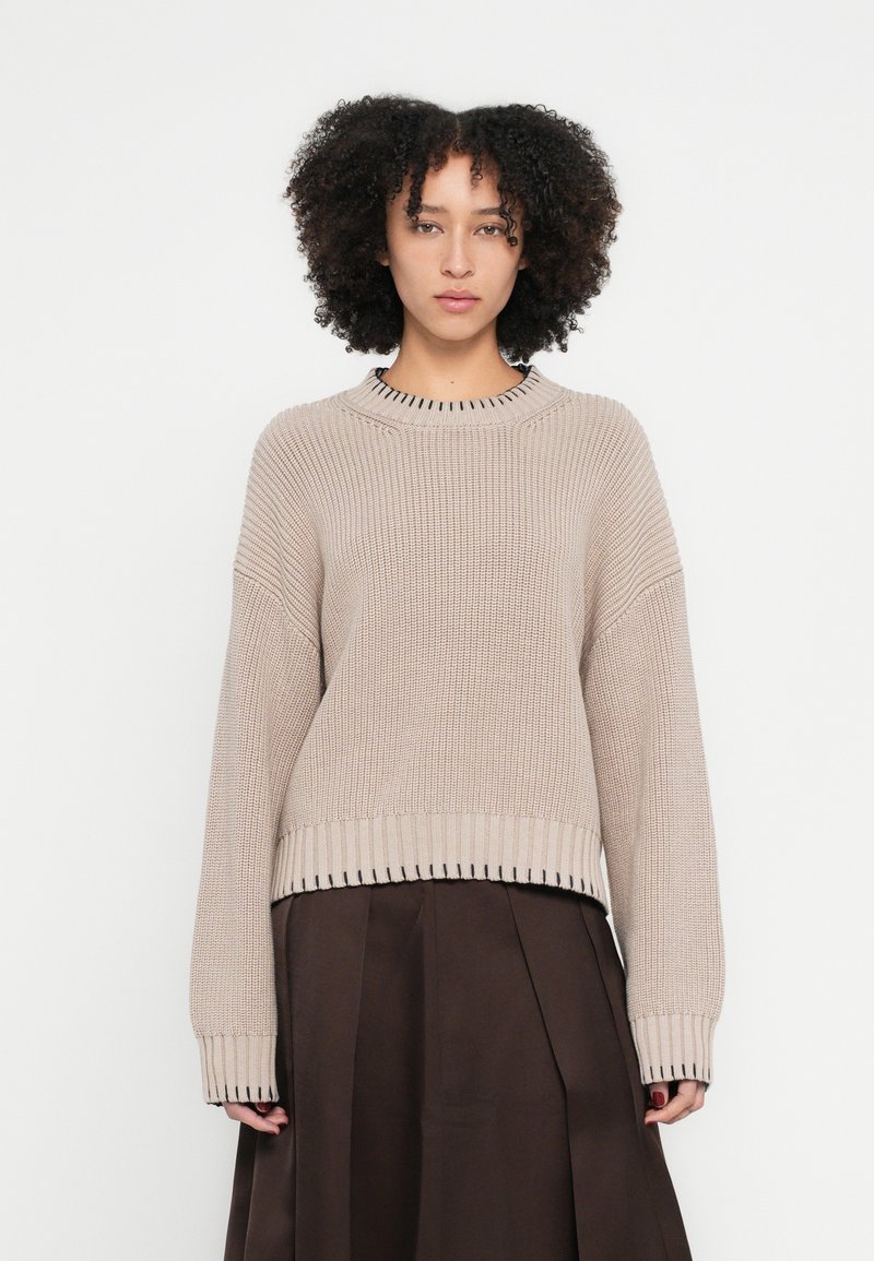 Bēša adīta džemperis ar nokarenām pleciem un ribotām aprocēm, ar melnām šuvēm apkārt apkaklei un apakšmalai. Valkāts pār brūnu plisirētu svārku.