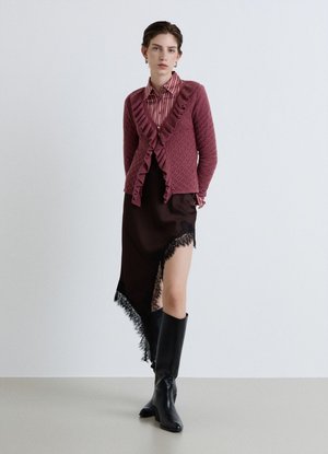 Donna che indossa un cardigan rosa con balze, camicia a righe, gonna nera asimmetrica bordata di pizzo e stivali neri al ginocchio, in piedi contro un muro bianco.