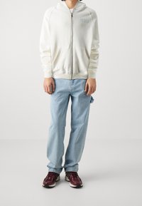 Felpa bianca con chiusura a zip e tasca a marsupio, abbinata a jeans azzurri e sneakers color bordeaux. La felpa presenta del testo ricamato sul petto.