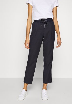 Person wearing cropped black drawstring pants, white short-sleeve shirt, and white sneakers, standing against a plain light background.