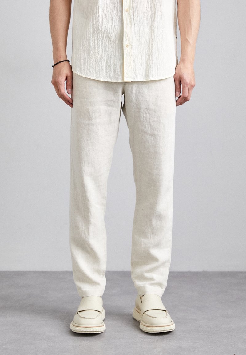 Jasnobeżowe lniane spodnie o luźnym kroju, zestawione z kremowymi mokasynami. Oba elementy garderoby cechują się gładką fakturą i minimalistycznym wzornictwem.