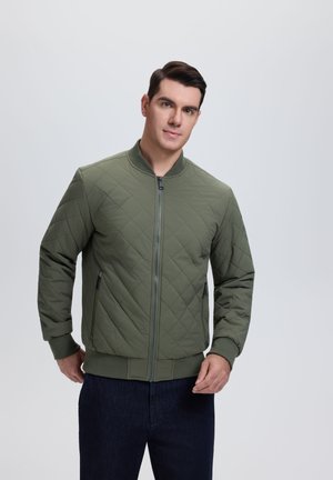 Man draagt een groene doorgestikte bomberjack met een ritssluiting aan de voorkant en donkere broek, staand tegen een lichtgrijze achtergrond.