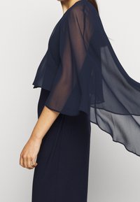 Marinblå klänning med genomskinliga fladderärmar och en figurnära silhuett. Tyget är slätt med en mjuk drapering som framhäver armarna.