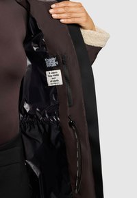 Giacca marrone con colletto in pelliccia, tasca interna con zip e etichette in tessuto. La fodera interna ha una texture liscia e un dettaglio con coulisse.