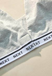 Šedý bavlnený bralet má dizajn v tvare „V“ s bielym elastickým pásom, na ktorom je v čiernej farbe napísané "NEXT". Mäkká textúra s hladkým povrchom.