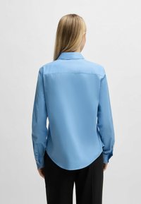 Camicia a maniche lunghe blu chiaro con colletto a punte, orlo curvo e polsini con bottoni. Tessuto liscio, con vestibilità rilassata e una leggera lucentezza.