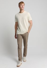 Lichtbeige crewneck t-shirt gecombineerd met slim-fit bruine broek, afgemaakt met witte sneakers met lichtbruine zolen.