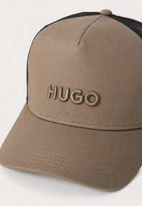 Căciulă de baseball bej cu față structurată, spate din plasă neagră și logo-ul brodat "HUGO" într-o nuanță mai deschisă. Detalii cusute pe bor.