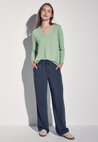 Suéter verde claro de cuello en V, de corte holgado, combinado con pantalones azul marino con cordón. El modelo lleva zapatos beige, mostrando un look casual y cómodo.