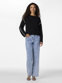 Woman with curly hair wearing a black sweater with white stitching, light blue straight jeans, and patterned open-toe sandals, standing against white background.