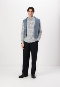 Cămașă cu mânecă lungă, cu un model albastru deschis, pantaloni bleumarin și un pulover albastru pus pe umeri; încălțările negre completează ținuta.