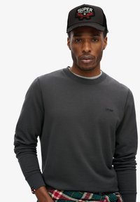 Sudadera gris de manga larga con cuello redondo, que presenta un sutil logotipo de la marca. Gorra de béisbol negra con un colorido parche bordado.