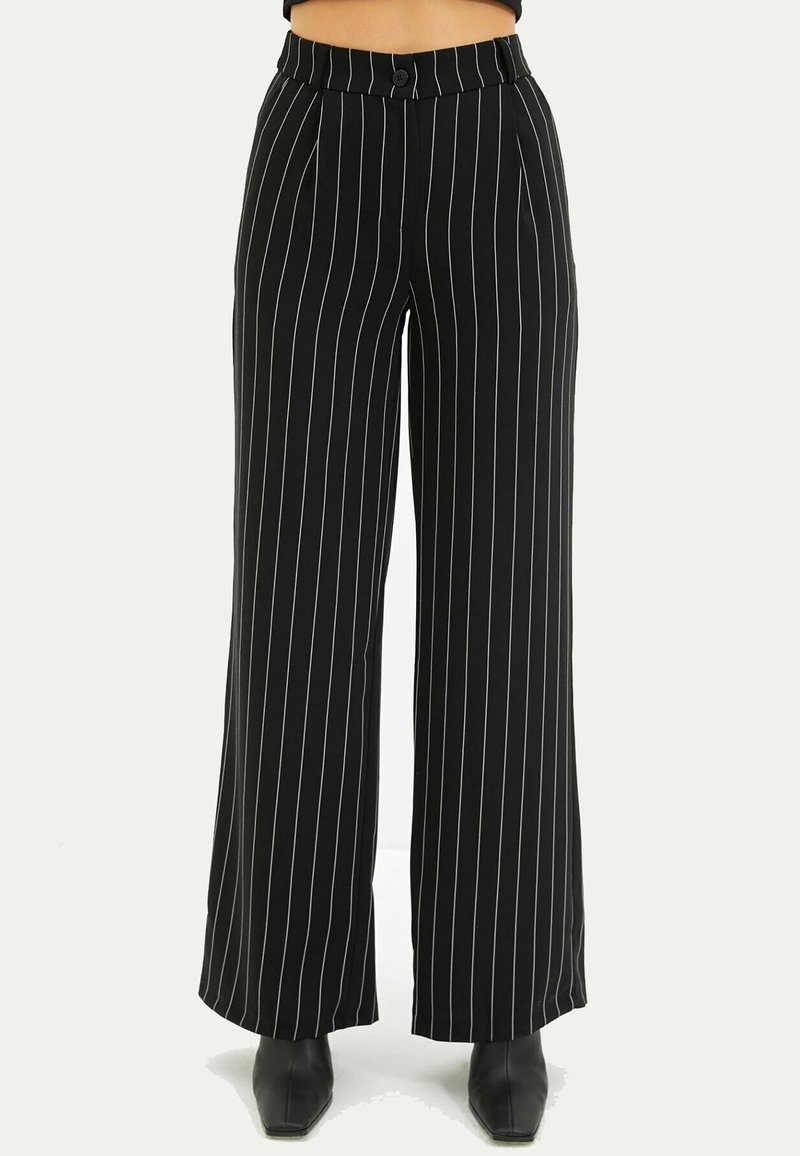 Pantalon noir taille haute à jambes larges avec fines rayures verticales blanches, fermeture à boutons devant, porté avec des bottes noires à bout carré.