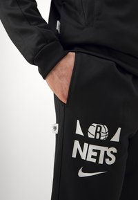 Pantaloni sportivi neri con una texture liscia, caratterizzati dal logo bianco "NETS" e dal baffo Nike. Tasche laterali e etichette brandizzate visibili.
